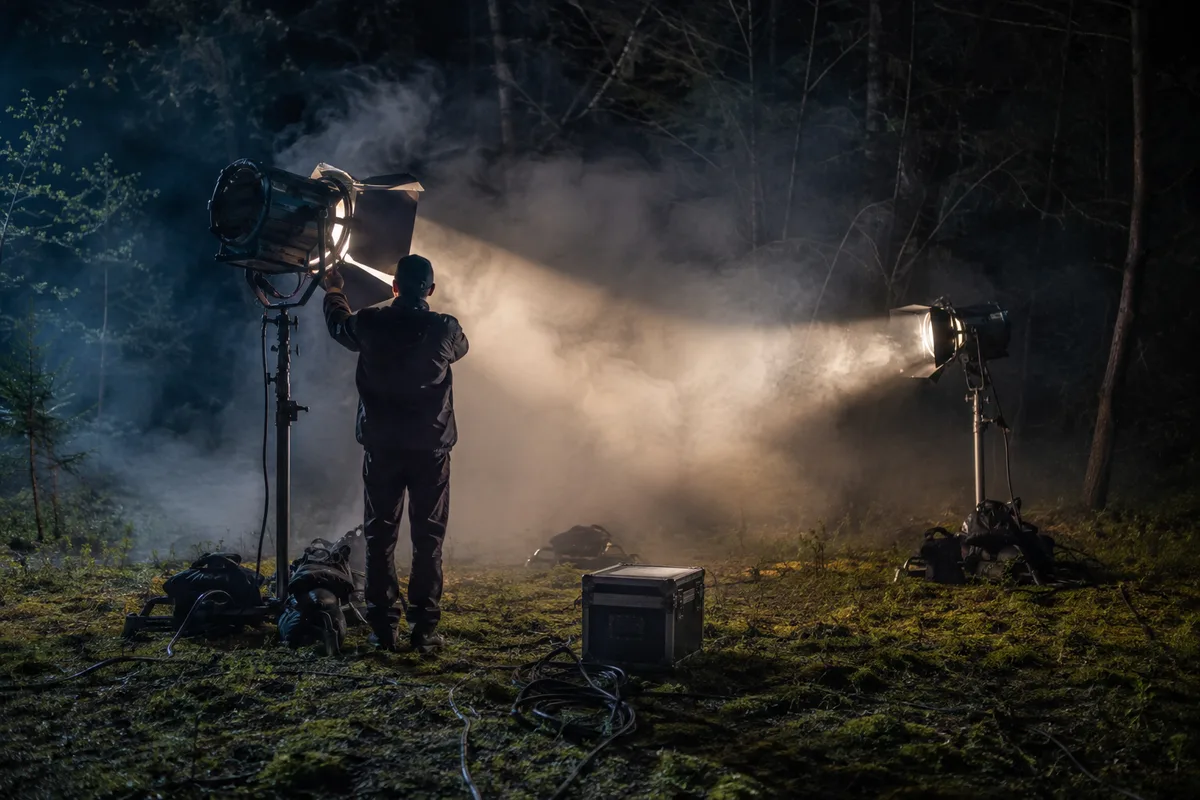 Plateau de tournage nocturne en forêt, projecteurs sur pieds et câbles au sol, halo de brume artificielle